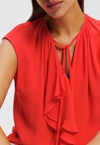 Blusa roja de tejido ligero con un frente con volantes, detalle de escote en forma de llave y mangas de capucha, textura suave y color vibrante.