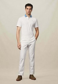 Weißes Poloshirt mit hellblauem Kragen, kurzen Ärmeln und kleinem Logo. Kombiniert mit weißen Hosen und braunen Turnschuhen. Einfaches Design.