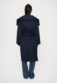 Long manteau bleu marine avec un grand col, taille ceinturée et coupe droite. Fabriqué dans un tissu texturé avec une finition lisse.