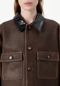 Brūna ādas jaka ar melnu spīdīgu apkakli, divām priekšējām kabatām un apaļām pogām. Mīksta tekstūra un kontrastējoši materiāli.