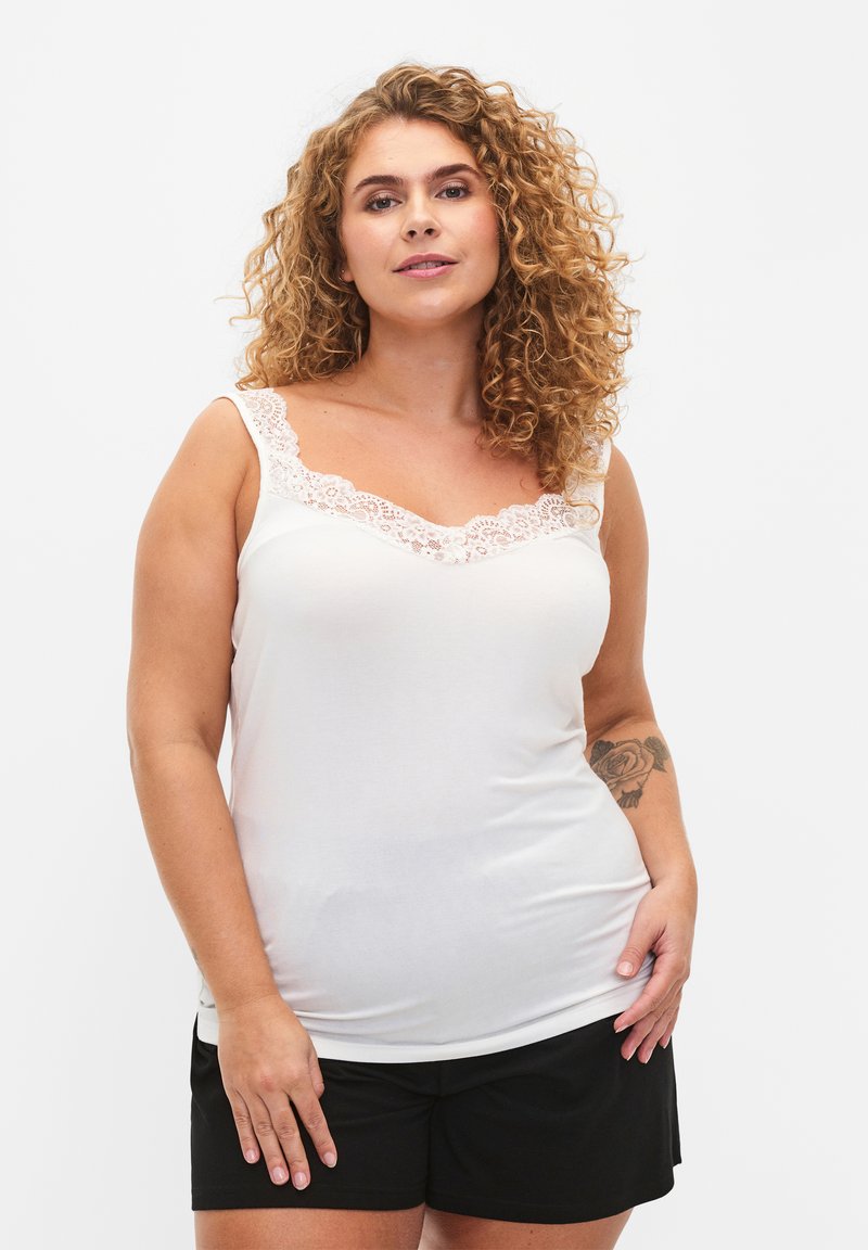 Débardeur blanc avec dentelle au niveau du col, en tissu doux, coupe slim, associé avec un short noir. Cheveux bouclés et tatouage floral visible sur le bras.