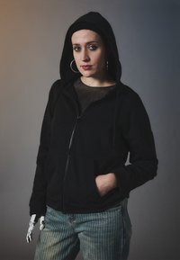 Crna zip-up dukserica s kapuljačom, nošena preko prozirne bluze. Manekenka nosi plave prugaste traperice i ima protezu na ruci.