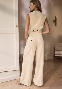 Top bej fără mâneci, cu un design ajustat și mici butoane negre la talie, asociat cu pantaloni cu picioare largi, tot bej, care au buzunare pe spate.