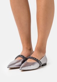 Piernas con zapatos planos plateados metálicos puntiagudos con gruesas correas de cadena negra metálica en la parte superior, sobre un suelo blanco.