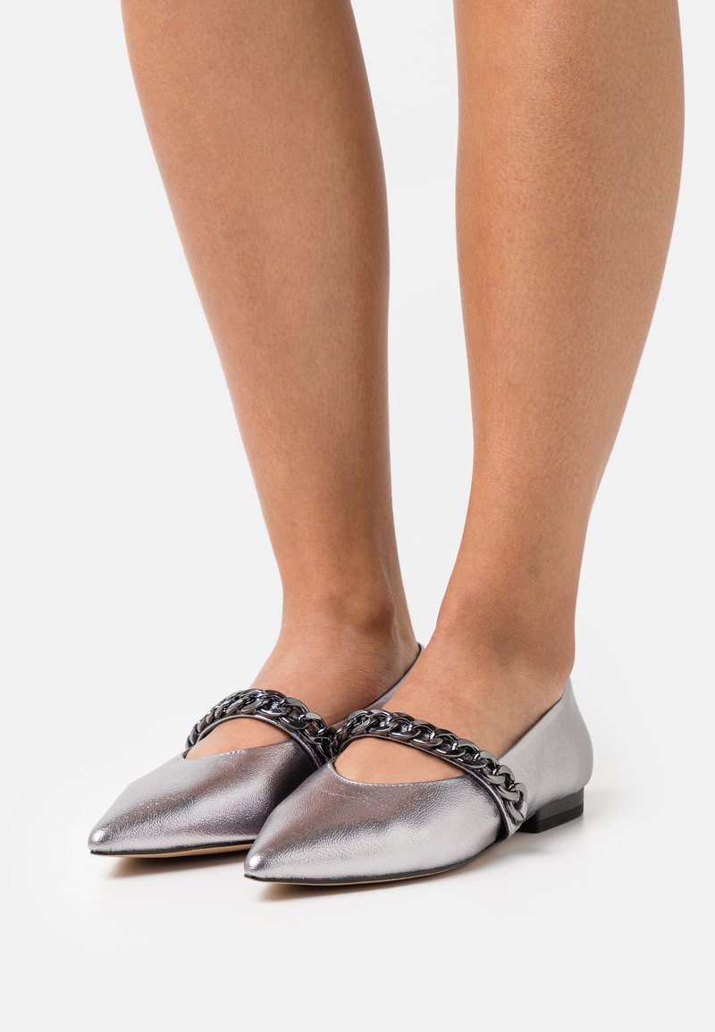 Piernas con zapatos planos plateados metálicos puntiagudos con gruesas correas de cadena negra metálica en la parte superior, sobre un suelo blanco.