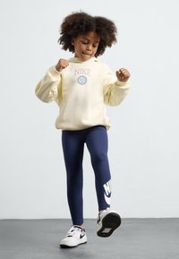 Dzeltena kapuce ar "Nike" logo, tumši zili legingi ar lielu balto "Nike" zvīņu un melnbalti sporta apavi. Mīksta auduma, brīva piegriezuma.