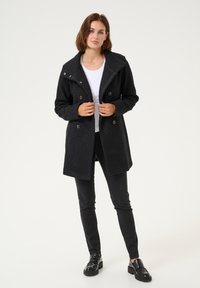 Zwarte dubbelgestikte jas met een gestructureerd oppervlak, grote kraag en knopenaccenten. Gedragen over een witte top en gecombineerd met zwarte broek en schoenen.