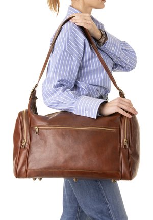 Donna con camicia a righe blu che porta alla spalla una grande borsa da viaggio in pelle marrone con molteplici tasche con cerniera.