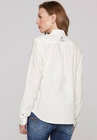 Camicia bianca a maniche lunghe con una piega posteriore e un dettaglio di ricamo a forma di ancora. Realizzata in morbido tessuto di cotone, presenta polsini abbottonati e un orlo curvo.