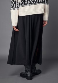 Jupe maxi noire avec fentes latérales, en tissu texturé. Associée à des bottines noires à boucles. Gros pull en tricot blanc avec des motifs noirs.