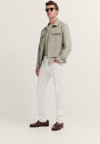 Light gray suede jacket with two chest pockets, green t-shirt, white denim jeans, and brown loafers; model wearing sunglasses.