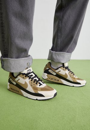 Person trägt beige, schwarze und braune Nike Air Max Sneaker mit hochgekrempelten grauen Jeans und steht auf grünem Boden.