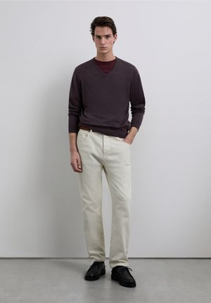 Dunkellila V-Ausschnitt-Pullover über einem kastanienbraunen Hemd, kombiniert mit cremefarbenen Hosen und schwarzen Schuhen, stehend vor einem neutralen Hintergrund.