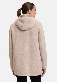 Beige Fleece-Mantel mit strukturierter Oberfläche, ausgestattet mit einer Kapuze und einem knopffreien Design, das bis zur Mitte der Oberschenkel reicht. Schwarze Hose darunter getragen.