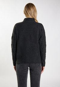 Mørkegrå strikket sweater med høj krave, der har kabelstrikkede detaljer på ærmerne og en ribbet kant. Simpel, struktureret design.