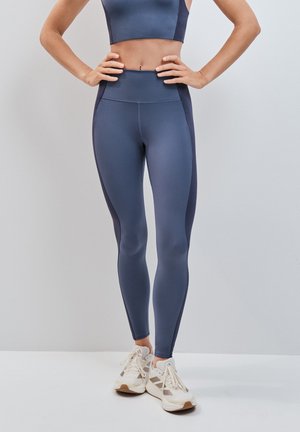 Person trägt hoch taillierte blaue Leggings und passendes Crop-Top, steht mit Händen in der Hüfte und weißen Sportschuhen vor einfarbigem Hintergrund.