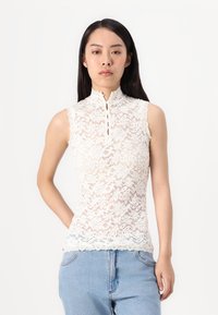 Čipkast bez rukava u off-white boji s visokim izrezom, s cvjetnim uzorcima i detaljem dugmeta na prednjem dijelu. Valoviti rub.