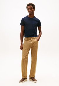 Marineblå bomuld t-shirt med korte ærmer, kombineret med tan farvede ribbukser og brune sneakers. Modellen poserer med den ene hånd i lommen.