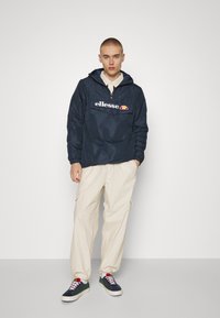 Anorak blu navy con cappuccio, una tasca frontale e logo; pantaloni cargo crema; sneakers verdi e blu navy con lacci rosa e suole bianche.