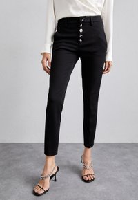 Schwarze, hoch taillierte Hose mit glatter Textur, fünf Knöpfen vorne und schmalem Schnitt. Kombiniert mit schwarzen Riemchen-High Heels mit silbernen Akzenten.