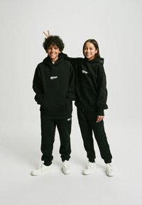 Dwoje dzieci nosi dopasowane czarne bluzy z kapturem i dresy wykonane z miękkiego materiału, mające kieszenie z przodu i akcenty w postaci logo, połączone z białymi sneakersami.