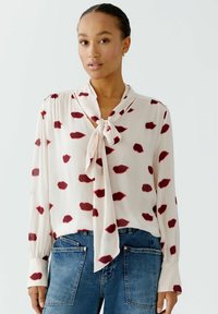 Weiße Bluse mit roten blütenblattähnlichen Mustern, langen Ärmeln und einer Schleifenkragen; kombiniert mit blauen Jeans. Glatte Stofftextur.