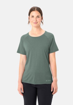 Frau mit zurückgebundenen braunen Haaren, die ein grünes Kurzarmshirt und schwarze Hosen trägt, vor einem einfachen hellen Hintergrund stehend.