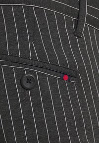 Gros plan sur la poche d'un costume gris foncé à fines rayures avec un bouton noir et un petit onglet en tissu avec un point rouge.