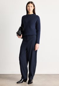 Γυναίκα με μπλε πουλόβερ και παντελόνι φαρδιάς γραμμής, κρατώντας μαύρο δερμάτινο τσαντάκι, στέκεται σε γκρι πάτωμα με φόντο λευκό τοίχο.