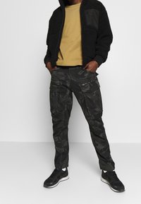 Man draagt een zwarte fleecejas, mosterdgele shirt, zwarte camouflage cargobroek met ritssluiting en zwarte sneakers, staat met handen in de zakken.