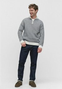 Man som bär en gråmönstrad kvartziptröja, mörkblå jeans med uppvikta benslut och bruna mockaskor, leende och stående.