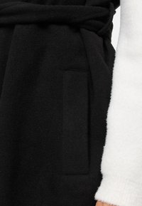 Robe noir en mélange de laine avec une finition texturée, une taille à nouer et une poche latérale ; une manche en polaire blanche est visible.