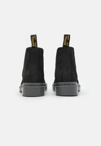 Bottines en suede noir avec des panneaux latéraux élastiques, semelles en caoutchouc gris plats, et étiquettes jaunes "Bouncing" à l'arrière pour un enfilage facile.