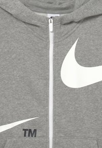 Grå zip-up hoodie i mjukt material, med en stor vit Nike swoosh-design och en liten svart "TM"-logotyp framtill.