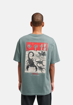Person wearing green T-shirt with a desert scene showing scorpions, cacti, and sun in red and black on the back.
