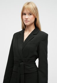 Μαύρο σακάκι με σχεδιασμό τύπου wrap και ζώνη για δέσιμο. Διαθέτει γιακά με εγκοπές και μία τσέπη στο στήθος. Λείο ύφασμα με δομημένη εφαρμογή.