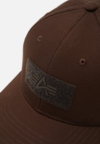 Șapcă de baseball maro cu panouri cusute și un patch texturat negru care spune "Alpha Industries" cu un logo stilizat "A".