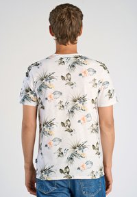 Valkoinen kukkakuosisella lyhythihaisella paidalla, jossa on vihreitä lehtiä ja persikanvärisiä kukkia. Paidassa on pyöreä kaula-aukko ja rento istuvuus.