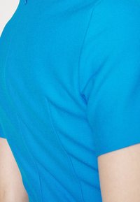 Modrý top s krátkými rukávy z hladké látky. Má přiléhavý střih s jemným prošitím a kulatý výstřih.