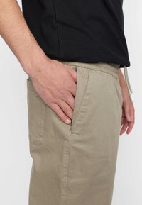 Beige katoenen broek met een zijzak met rits. Elastische tailleband met een verstelbaar trekkoord. Eenvoudig, functioneel ontwerp.