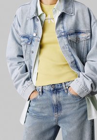 Ljusblå denimjacka över en gulribbad linne, med en vit skjorta som kikar ut. Höga ljusblå jeans. Avslappnad passform.