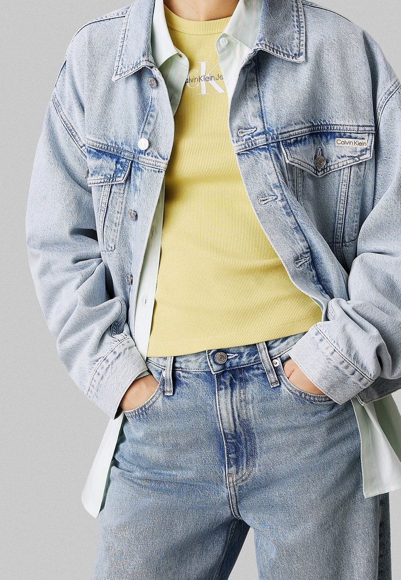 Ljusblå denimjacka över en gulribbad linne, med en vit skjorta som kikar ut. Höga ljusblå jeans. Avslappnad passform.