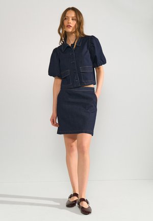 Femme porte une veste courte en denim bleu foncé avec des boutons et une jupe assortie, debout avec une main dans la poche et des chaussures plates marron.