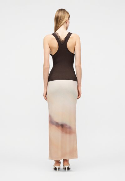 Γυναίκα με μακριά ξανθά μαλλιά, που φοράει μαύρο αμάνικο τοπ με λεπτομέρειες δαντέλας και μακριά μπεζ φούστα με σκούρο αφηρημένο σχέδιο, που στέκεται με την πλάτη.