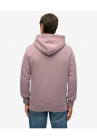 Sudadera con capucha en tejido púrpura claro, con cordones en la capucha, puños acanalados y un corte relajado, llevada sobre unos pantalones vaqueros de mezclilla azul.