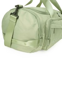 Bolsa de deporte de tela verde con forma redondeada, presenta cremalleras, correa ajustable y detalle del logo en el lateral. Textura suave, diseño minimalista.