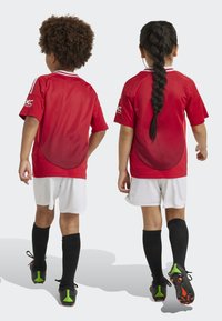 Des maillots de football rouges avec des accents blancs, associés à des shorts blancs et des chaussettes noires. Deux enfants, vus de dos, portent des chaussures de football noires.
