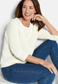 Frau lächelt, sitzt mit einem Arm auf dem Knie, trägt einen strukturierten weißen Pullover und Jeans in Blau vor einem schlichten Hintergrund.