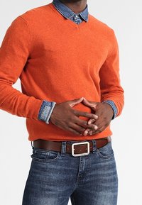 Oranget v-ringad tröja med en texturerad yta, lagd över en jeansskjorta. Mörkblå jeans med ett brunt bälte som har en silverfärgad spänne.