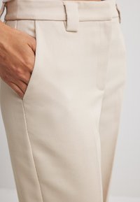 Beige ribbed trousers featuring a side pocket and a flat front. The fabric has a smooth texture with a tailored fit and belt loops.